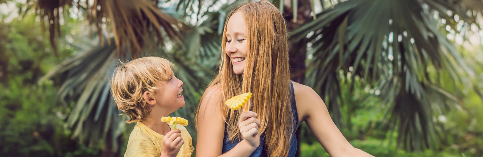 A smiling mother and son sitting outdoors in front of tropical palm trees, while holding pineapple slices on sticks